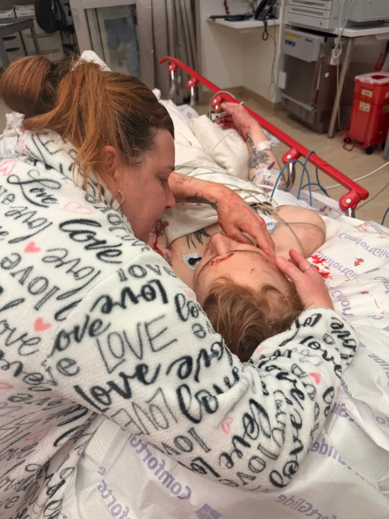 A woman in a "love" themed robe embraces a young man with visible injuries and medical equipment in a hospital bed.