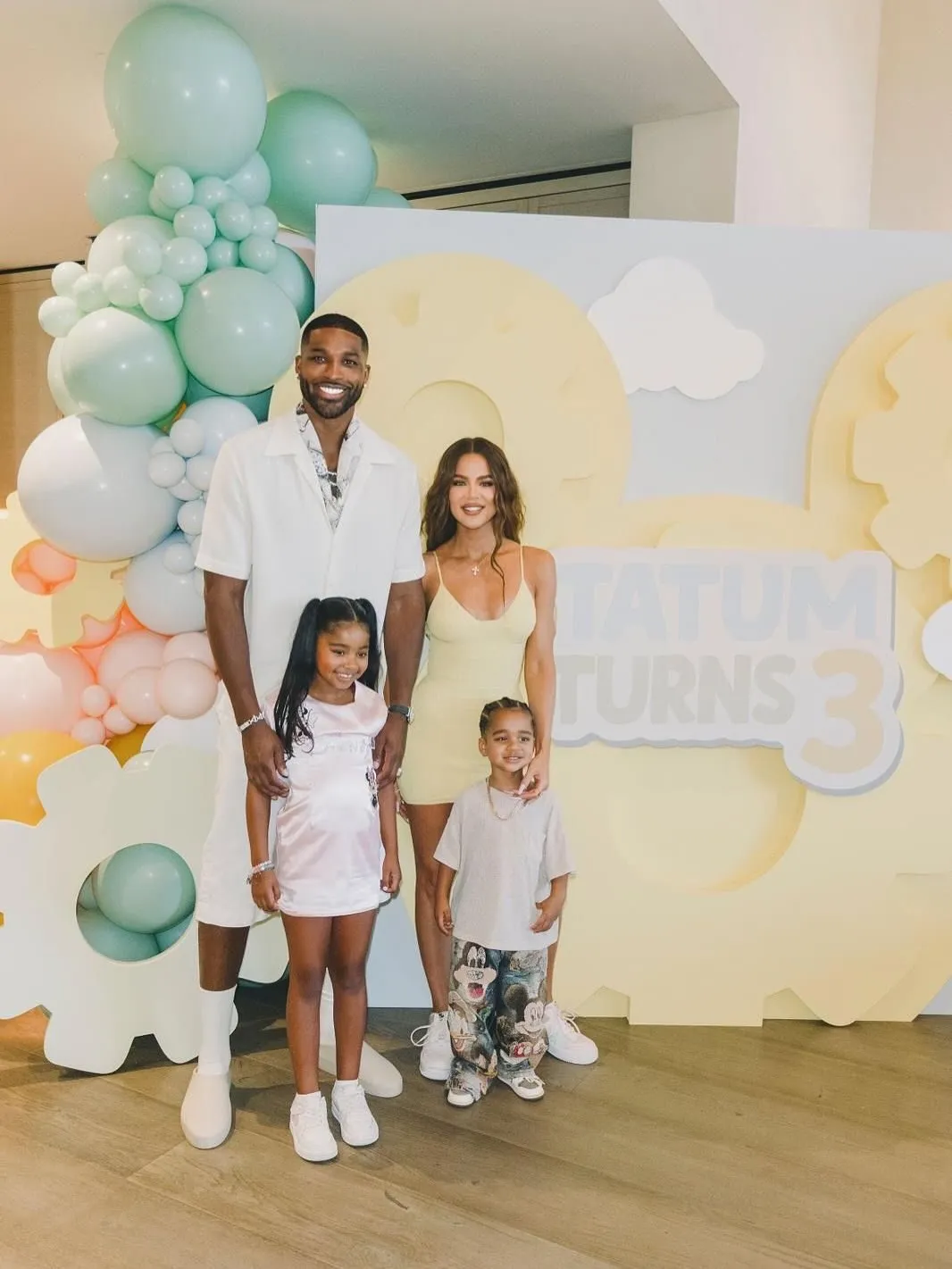 Tristan Thompson and Khloé Kardashian pose with their children, True and Tatum, in front of a light yellow and blue "Tatum Turns 3" birthday display.