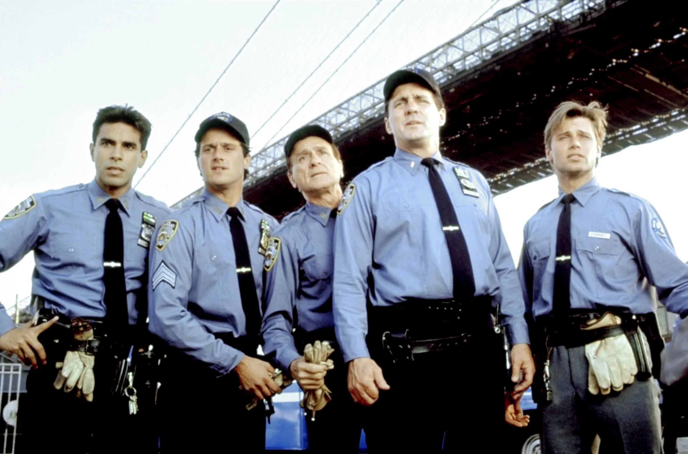 Cast photo of five actors dressed as police officers from the television series "True Blue," standing under the Brooklyn Bridge.