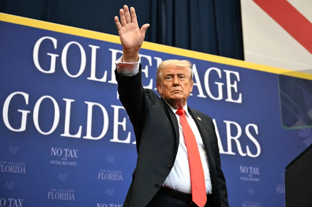 President Trump raises his hand while standing in front of a blue banner that reads "NO TAX ON SOCIAL SECURITY".