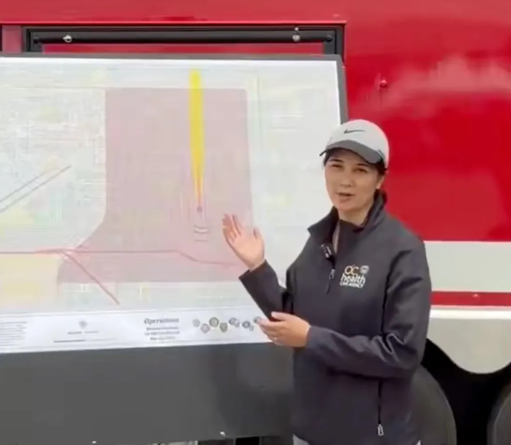 A woman wearing an "OC Health" jacket and a baseball cap gestures at a map of evacuation zones.