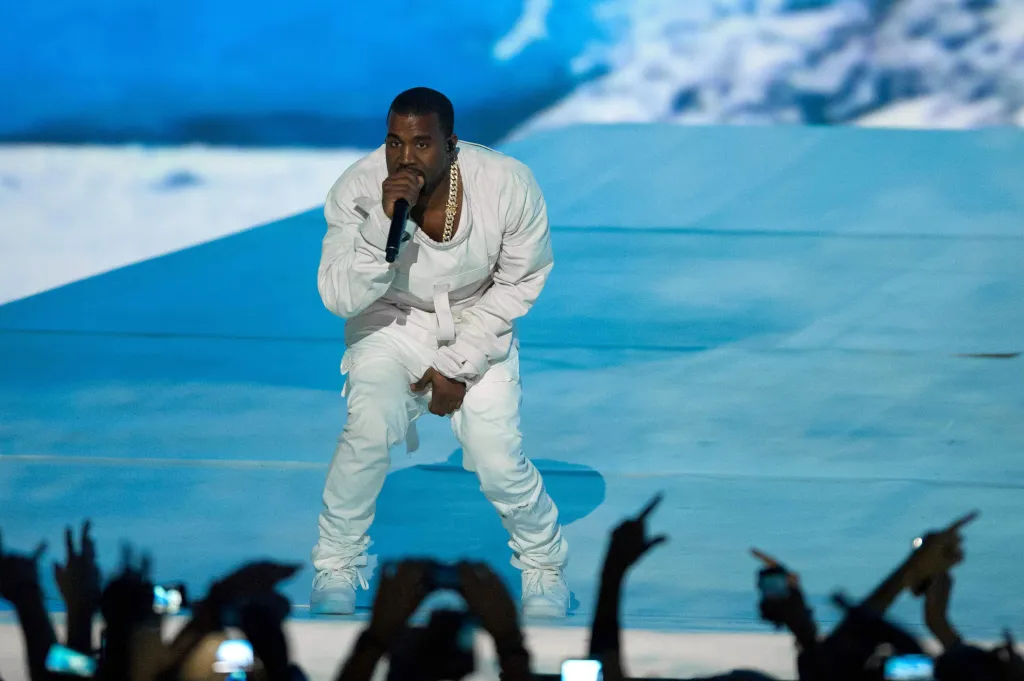 Kanye West performing on stage in a white outfit, holding a microphone.