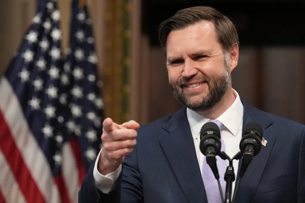 JD Vance, smiling and pointing, stands behind microphones with American flags blurred in the background.