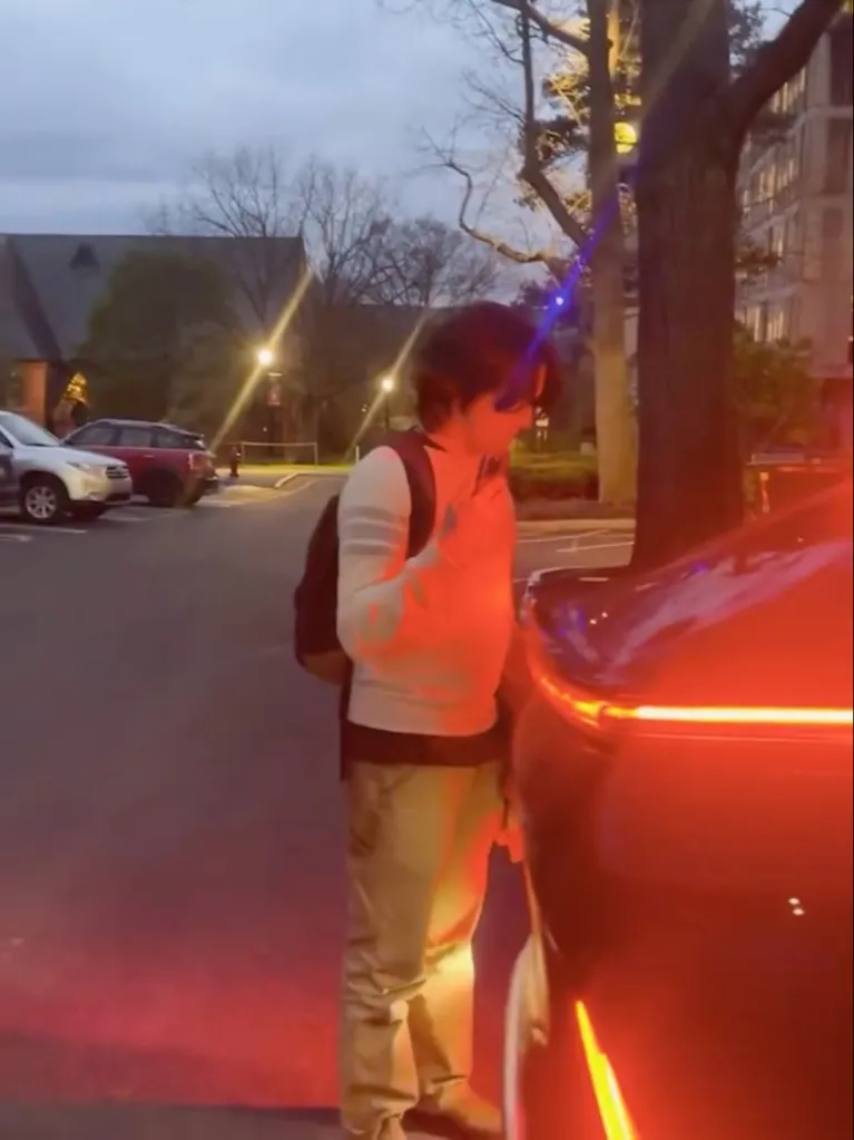 A student wearing a backpack stands next to a car with its brake lights illuminated, as if it is reversing.