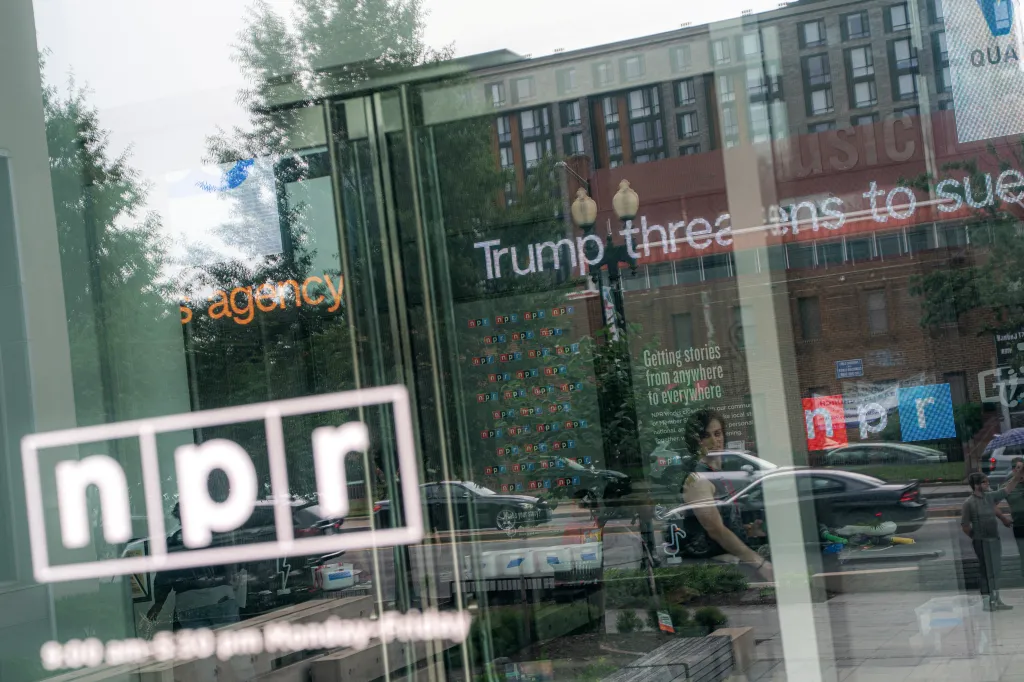 Reflection of the Washington D.C. NPR headquarters and a "Trump threatens to sue" news headline on a window.