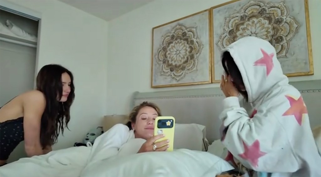 Alix Earle watching a video on a phone while laying in bed as two other women are also in the room.