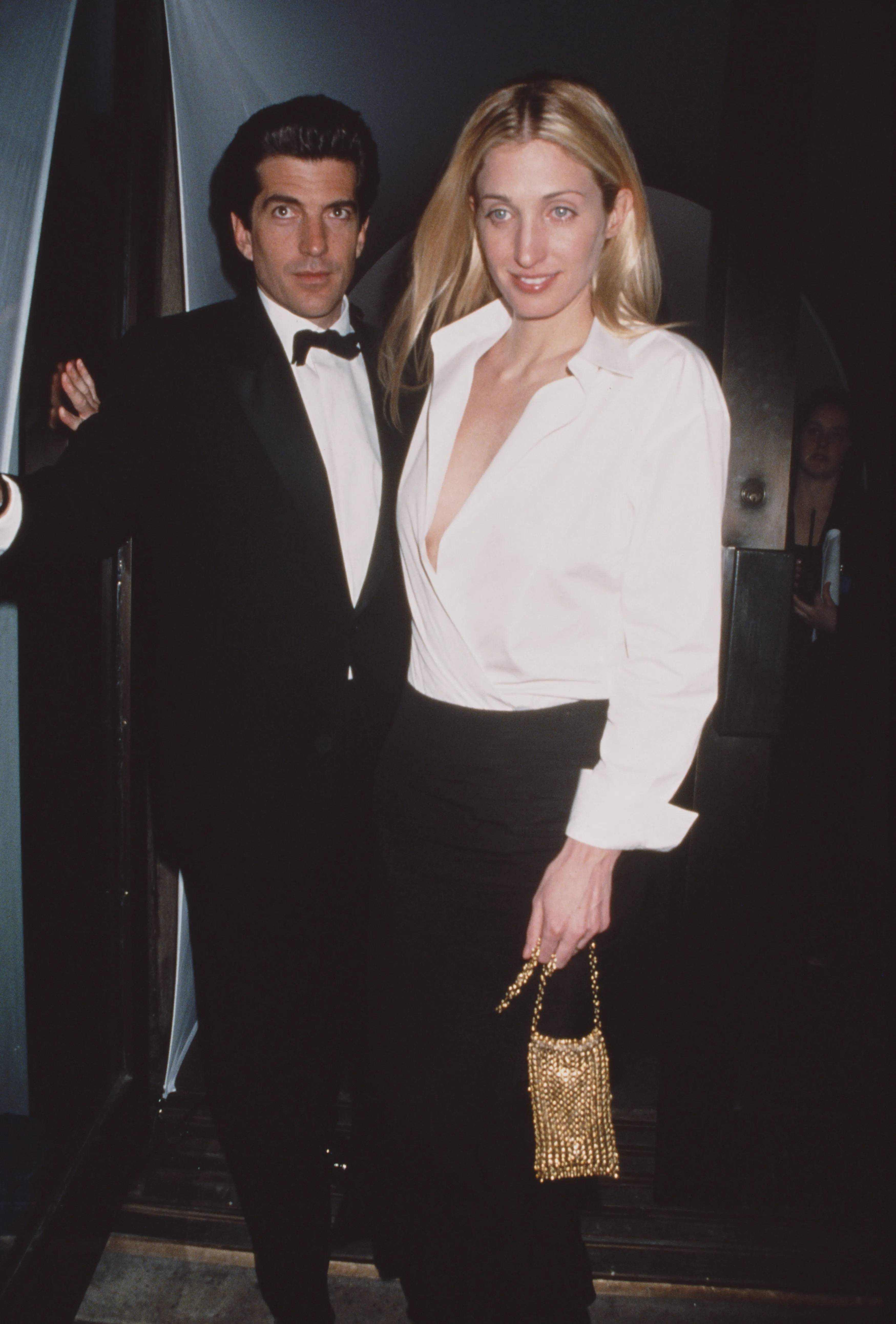 John F. Kennedy Jr. in a tuxedo with his wife, Carolyn Bessette-Kennedy, in a white shirt and black skirt.