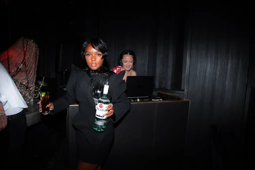 Janelle Monáe holding a Bacardi bottle and a drink, with a DJ in the background.