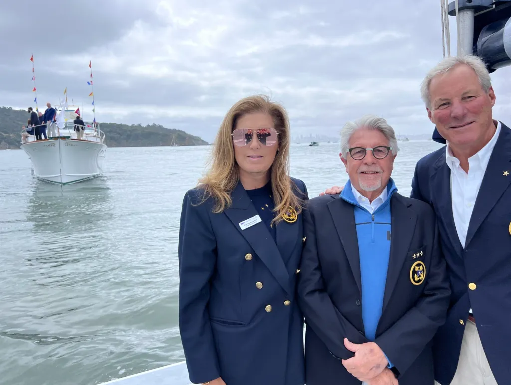 Marisa McArthur, wearing sunglasses and a navy blazer, stands on a boat with two men, also in blazers, with another boat in the background.