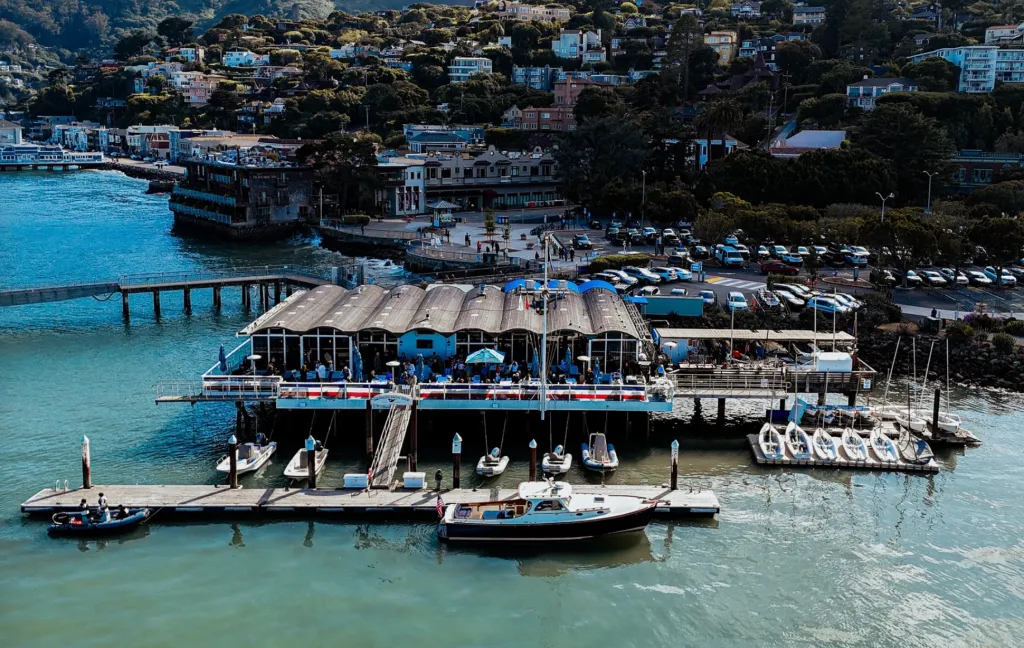 View of the Sausalito Yacht Club from above, featuring the club building on the water, docked boats, and a hillside town in the background.
