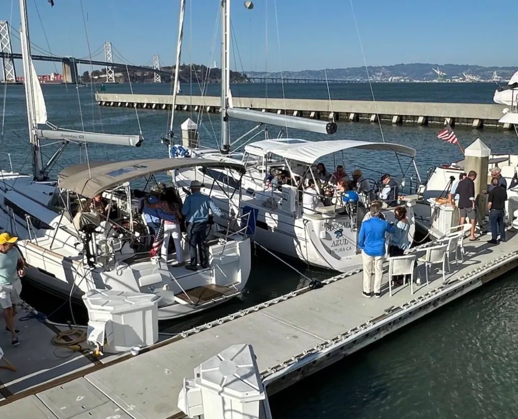 People gathered on sailboats docked at Sausalito Yacht Club, with a bridge in the background.