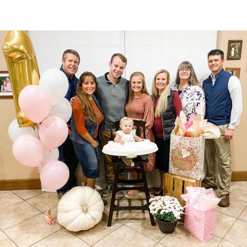 A baby in a high chair celebrating a birthday with seven adults and balloons.