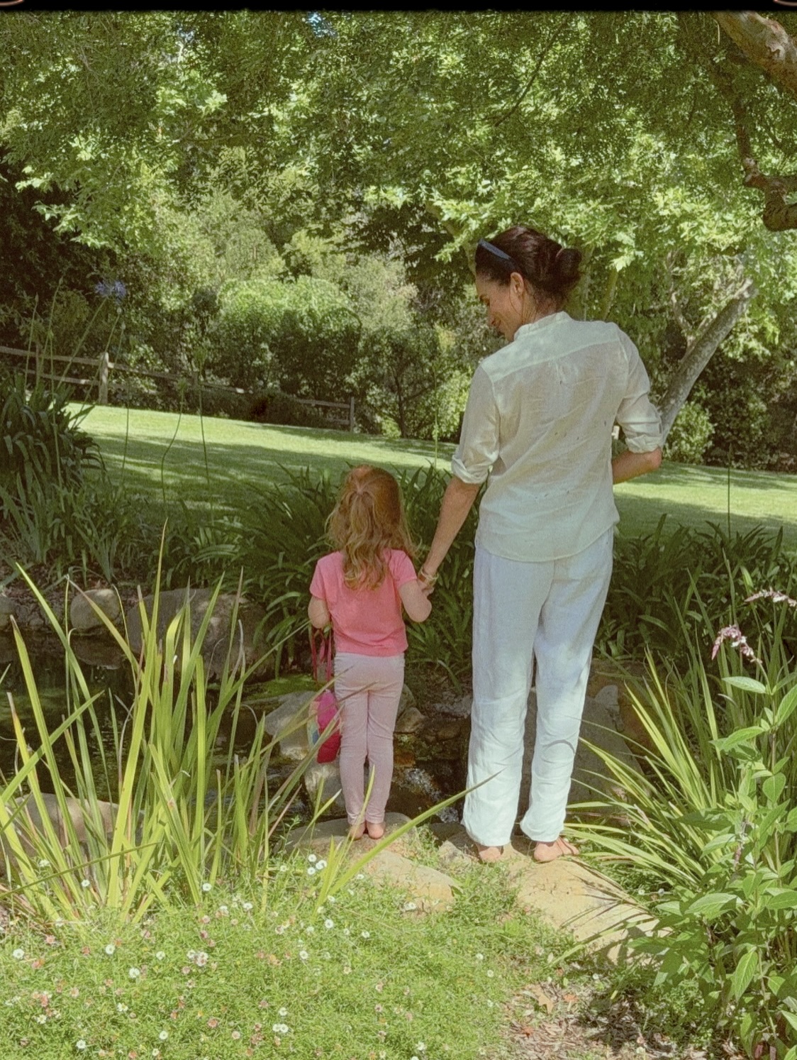 Meghan Markle holding hands with her daughter, whose red hair is visible, as they stand barefoot by a stream.