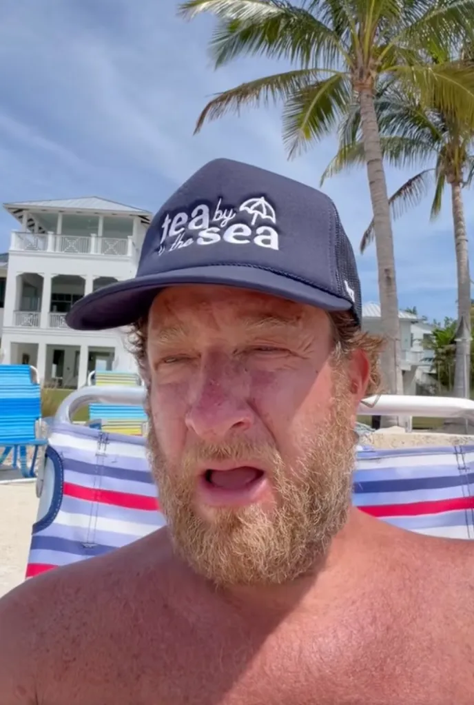 Dave Portnoy wearing a "Tea By The Sea" hat, sitting in a beach chair with his mouth open, on a beach with palm trees and a large white house in the background.