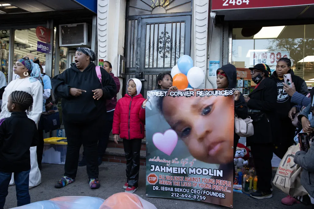 People gather on a sidewalk for a balloon release, holding a poster with a child's photo and the text