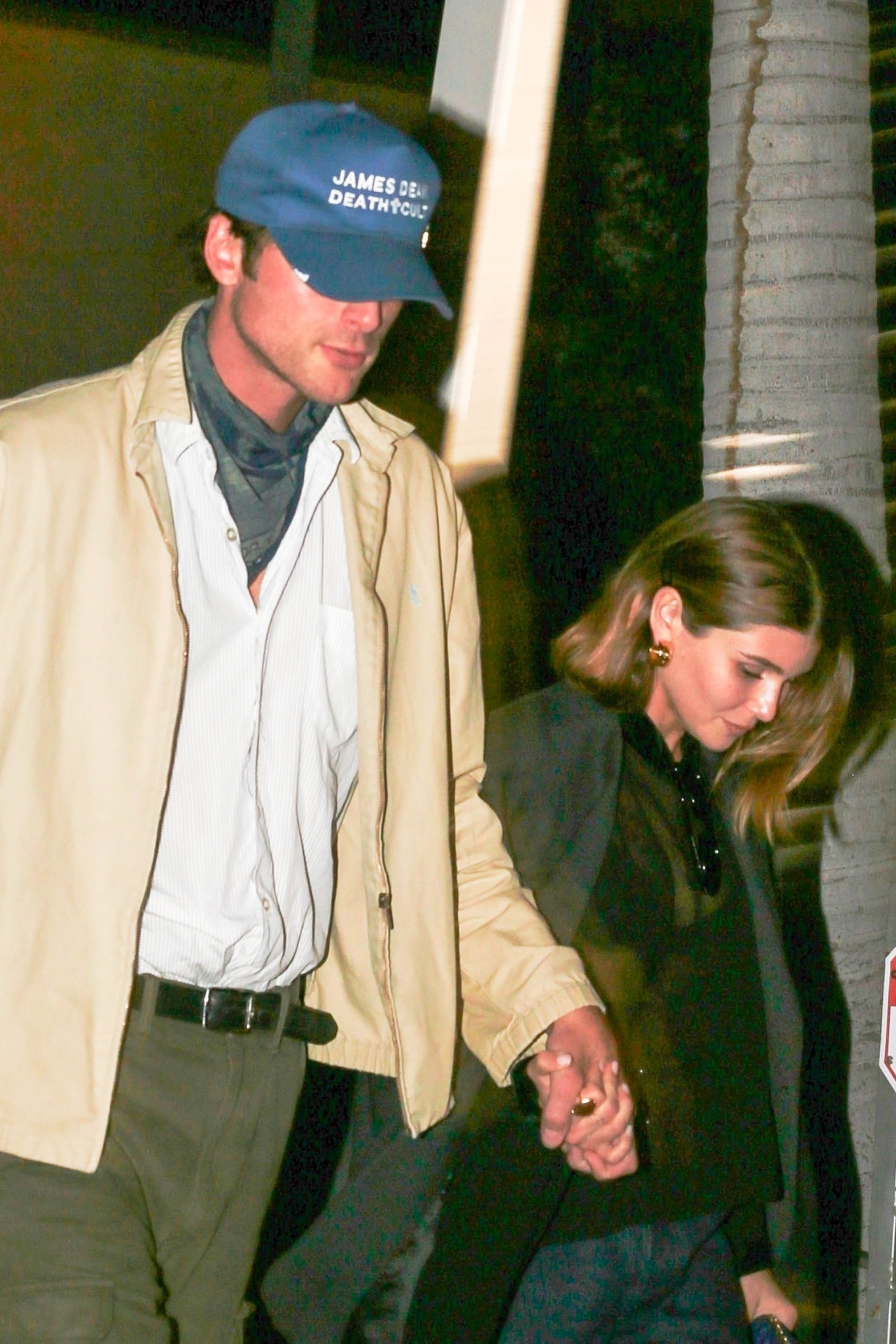 Jacob Elordi, wearing a blue "James Dean Death Cult" baseball cap and a tan jacket, holding hands with Olivia Jade.