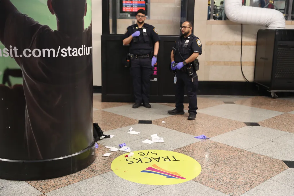 Two police officers stand near bloodied debris on the floor, including a yellow "TRACKS 5/6" sign.