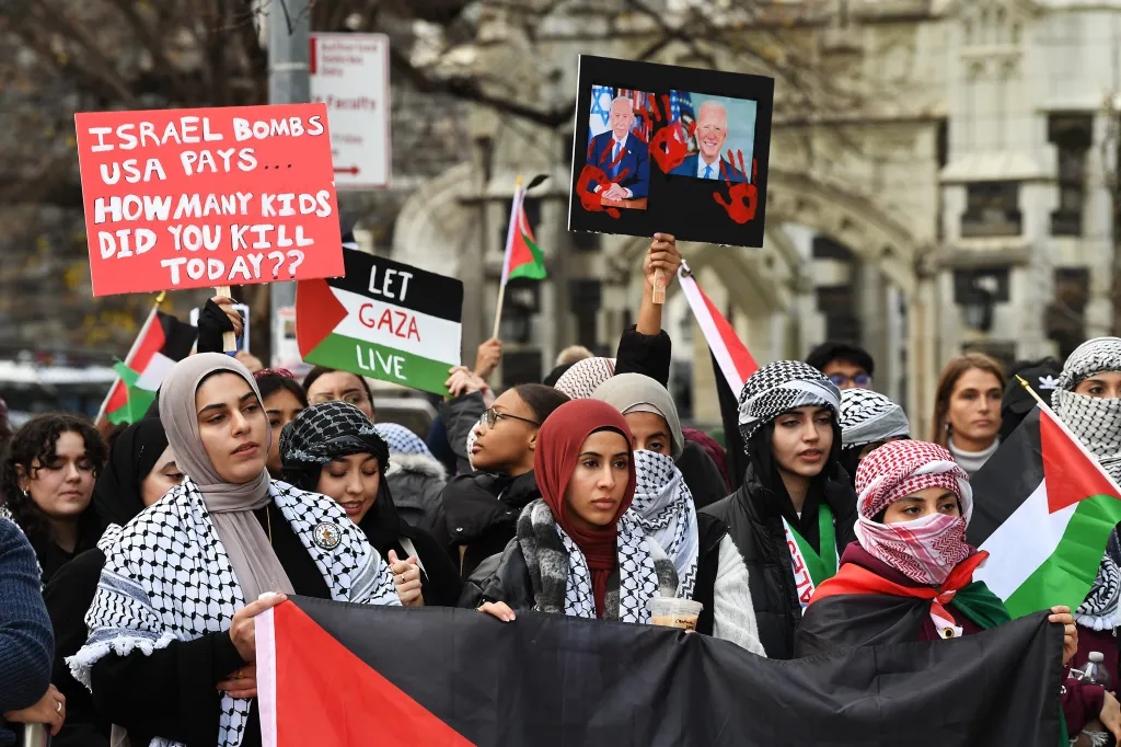 People protesting in support of Palestine, holding signs that read "Israel bombs USA pays... How many kids did you kill today??" and "Let Gaza Live", and a sign featuring images of President Biden and Prime Minister Netanyahu with bloody handprints.