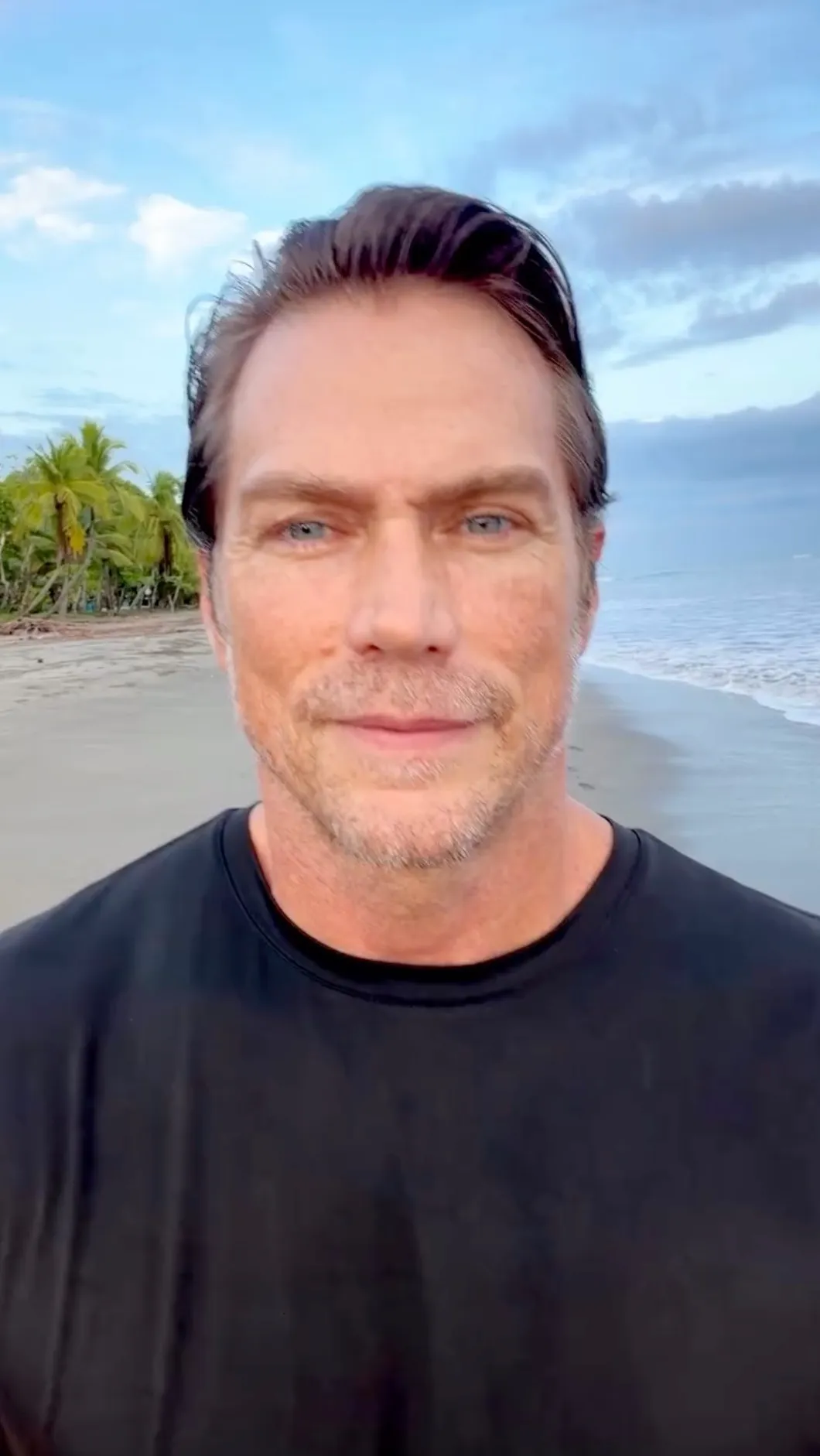 A man with blue eyes and a slight beard, wearing a black shirt, on a beach with palm trees and ocean in the background.