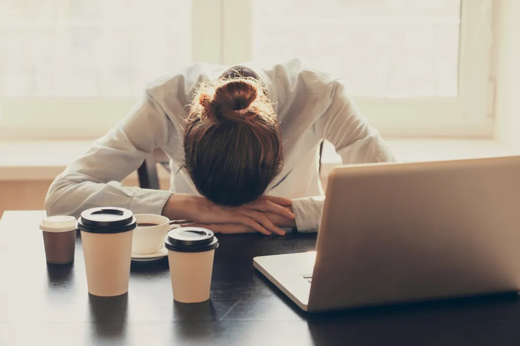 A tired woman with her head down on her arms at a desk, surrounded by several coffee cups and a laptop.
