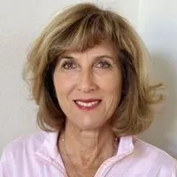 A woman with shoulder-length blonde hair, bangs, and a pink shirt smiles at the camera.
