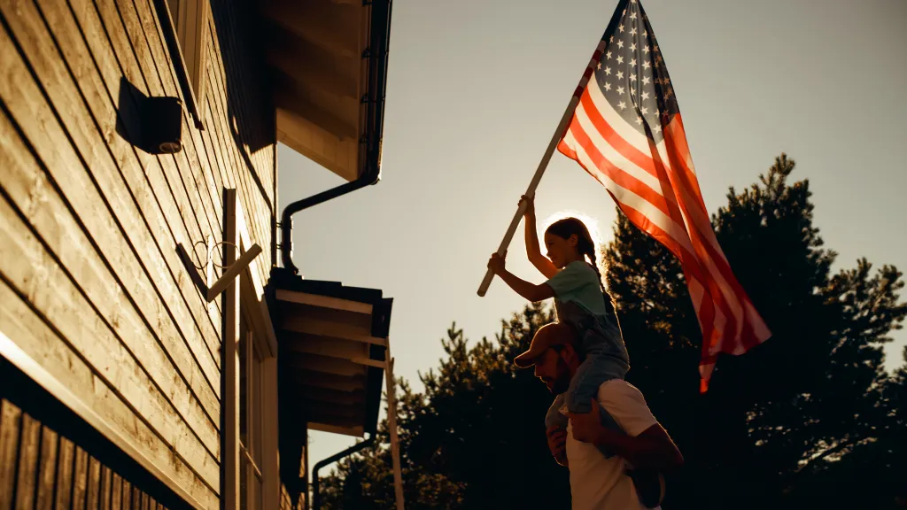 Father holding his daughter on his shoulders as she helps raise an American flag at their house.