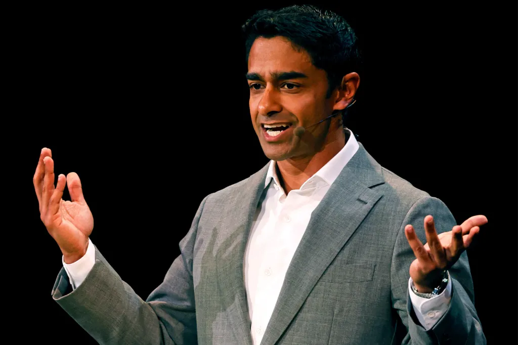 A man with dark hair in a gray suit and white shirt with a microphone visible on his cheek, speaking with his hands outstretched.