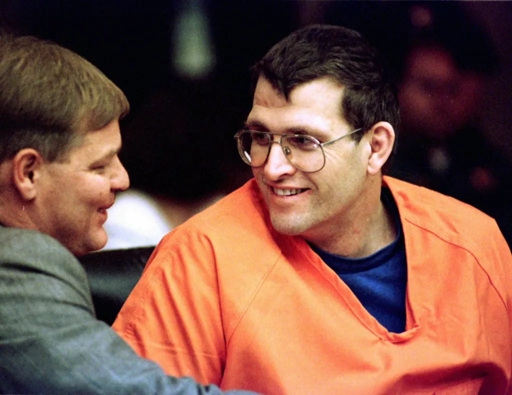 Convicted murderer Keith Jesperson, known as the "Happy Face Killer", in orange prison attire and glasses, smiling next to his attorney Thomas Phelan.