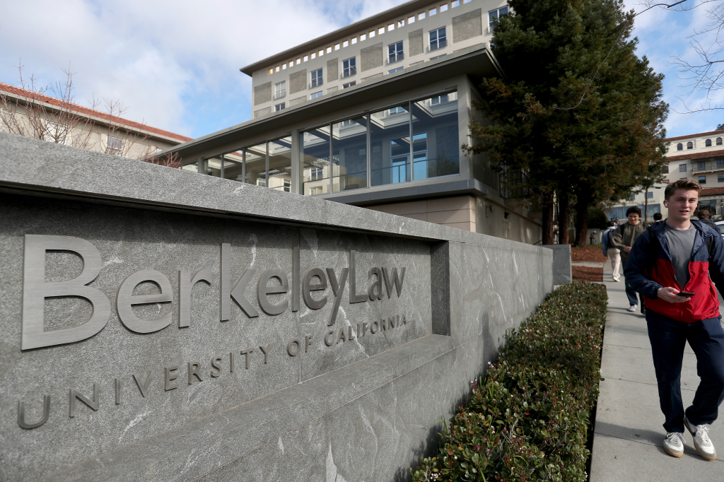 The "Berkeley Law" sign on a stone wall, with a student walking past and the university building in the background.
