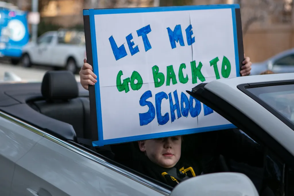 A boy holds a "Let Me Go Back To School" sign from the back seat of a convertible car at a rally.