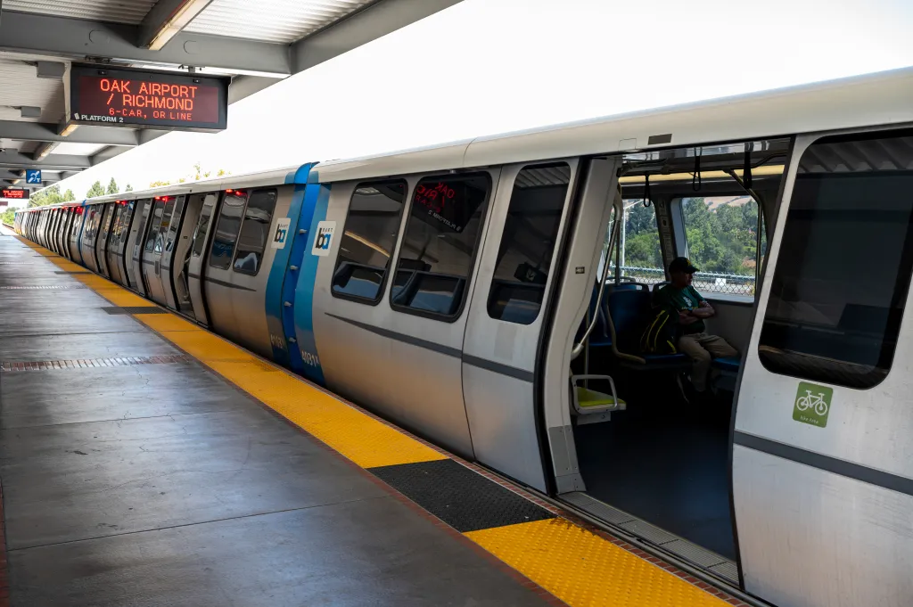 A BART train stopped at a station with a sign overhead displaying "OAK AIRPORT / RICHMOND."