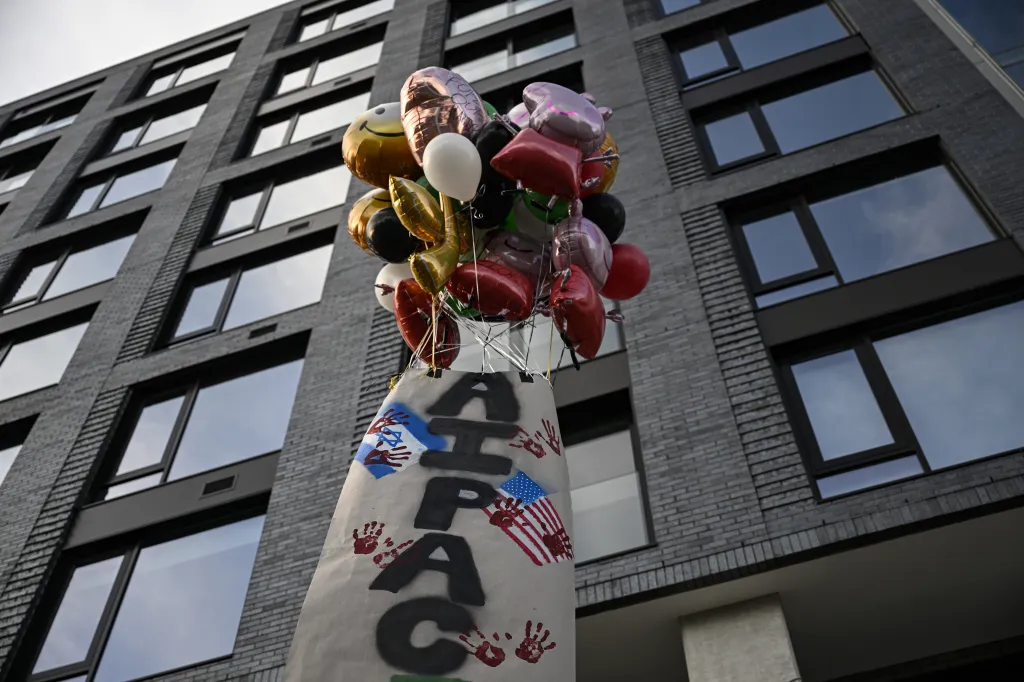 Protest sign with "AIPAC" and bloody handprints, flanked by Israeli and American flags, tied to balloons outside a building.