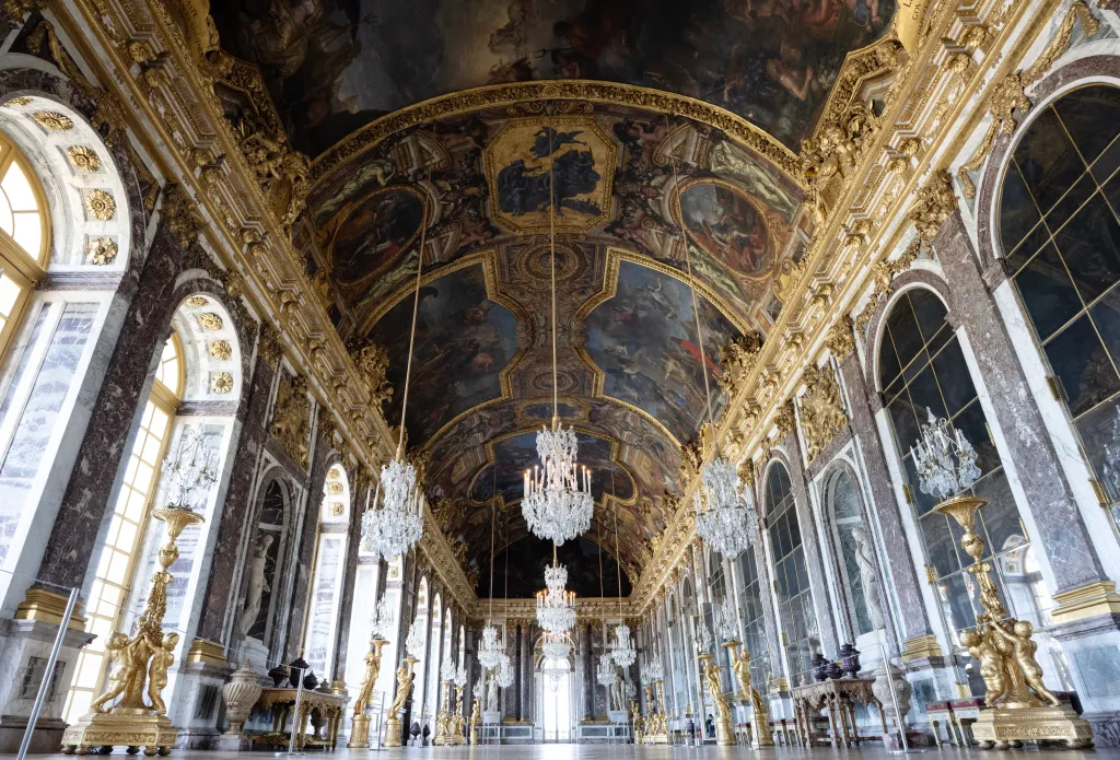 The Hall of Mirrors inside the Palace of Versailles, featuring ornate chandeliers, gilded decorations, and painted ceilings.