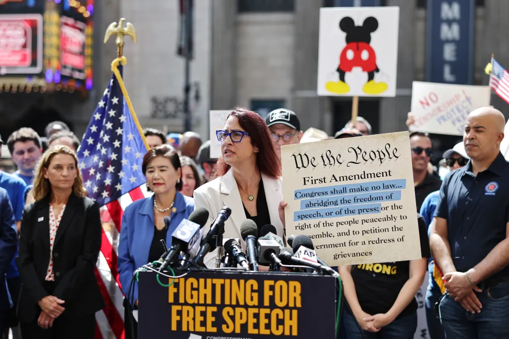 U.S. Rep. Laura Friedman speaks at a podium with microphones, holding a sign displaying the First Amendment, during a protest against the suspension of the "Jimmy Kimmel Live!" show.