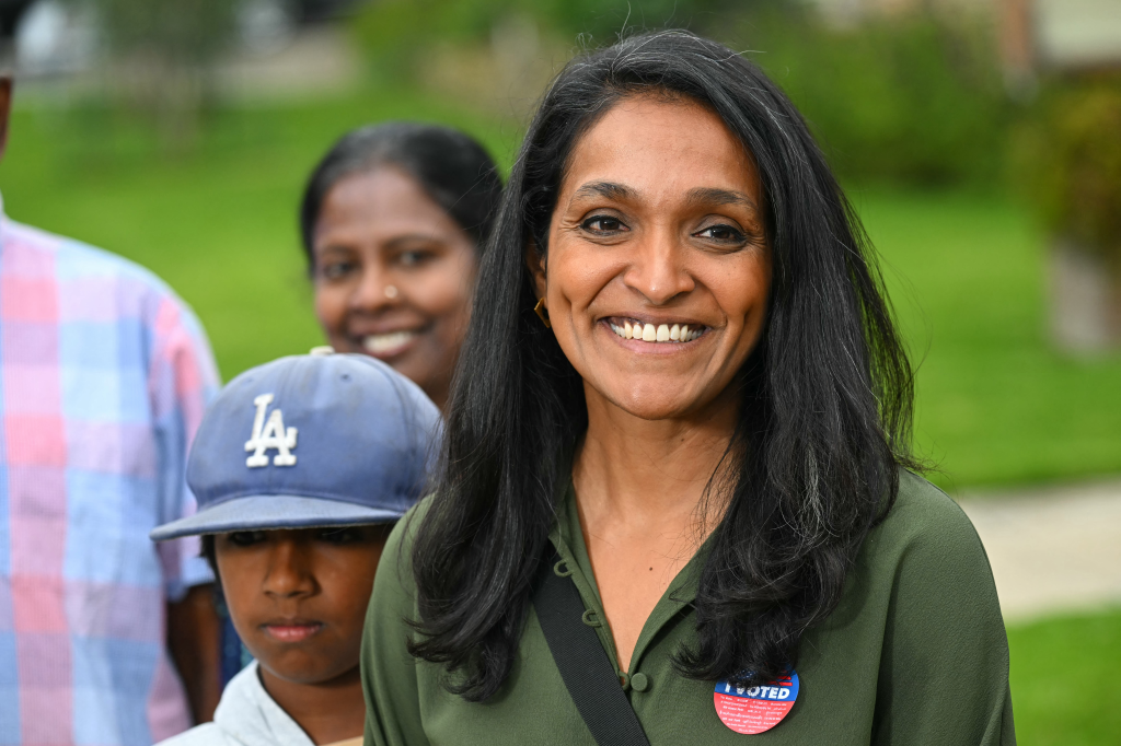 Los Angeles Councilmember Nithya Raman smiles, wearing an "I Voted" sticker, with a woman and boy in the background.