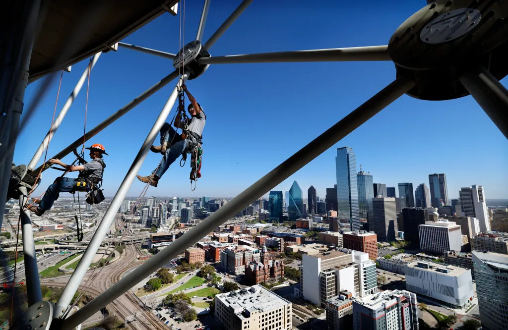Two technicians suspended by ropes adjust new LED lighting on the lattice shell of Reunion Tower, with the Dallas skyline visible behind them.