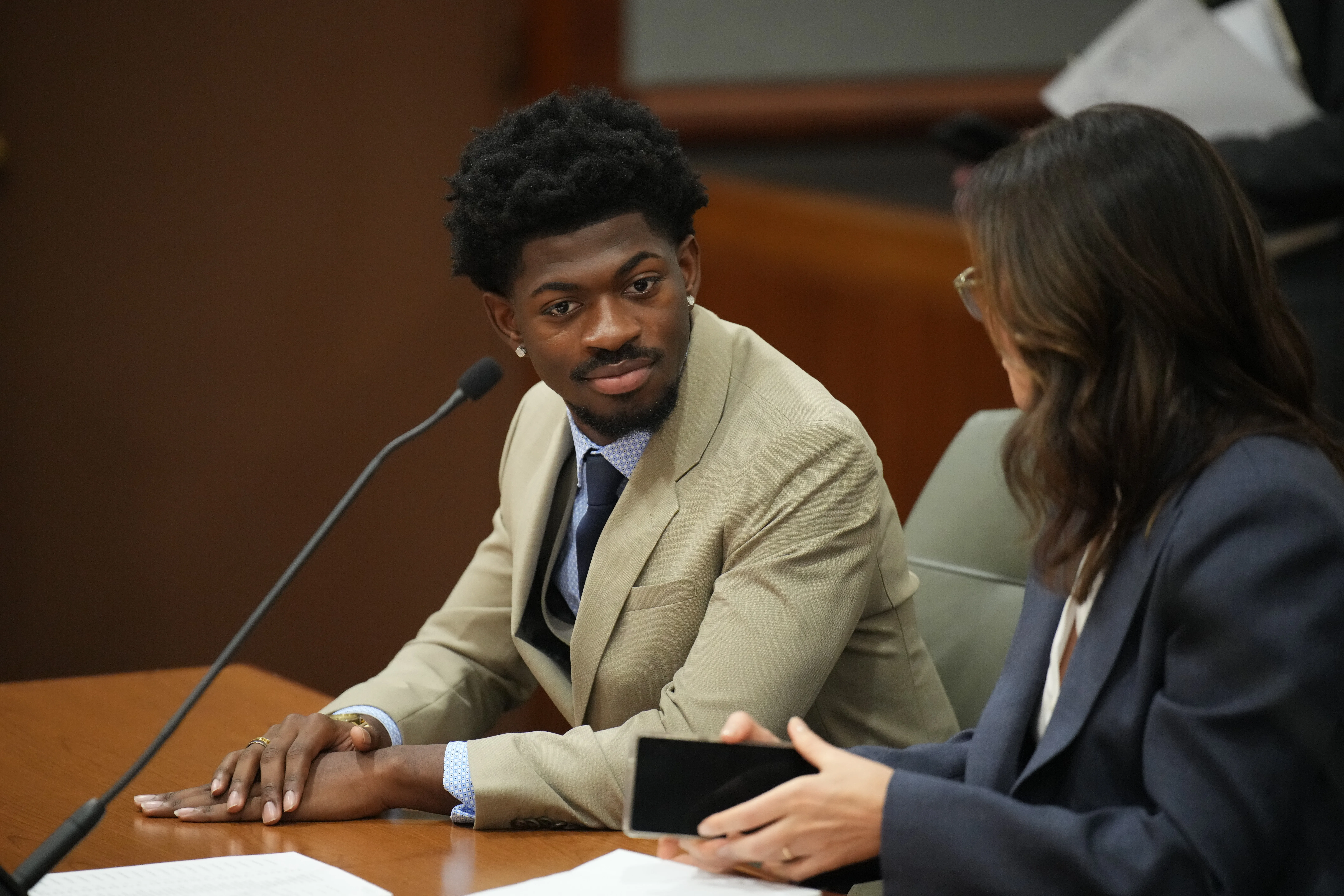 Lil Nas X in court, wearing a tan suit and looking towards a person across the table.