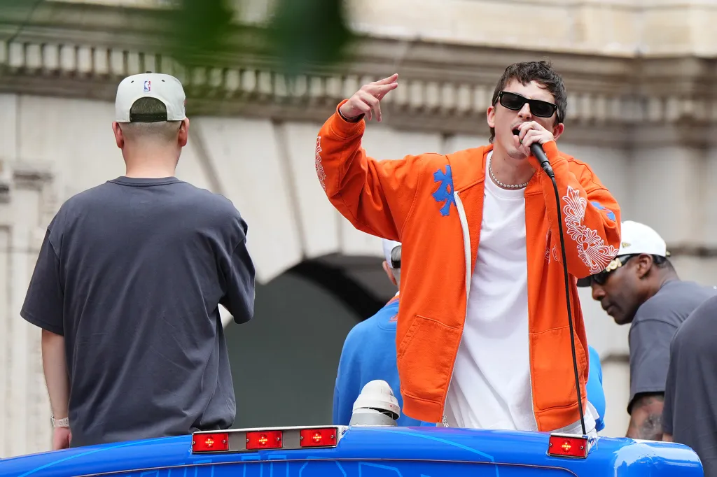 Timothée Chalamet singing into a microphone, wearing sunglasses and an orange jacket, on a parade float.