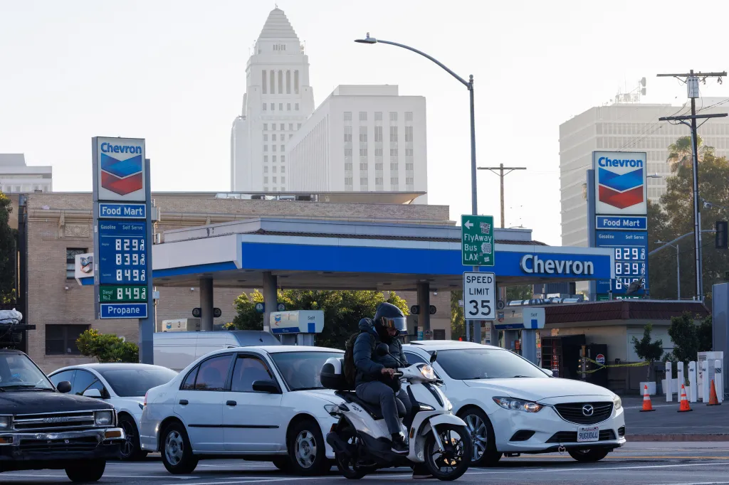 A person on a scooter rides past a Chevron gas station in Downtown Los Angeles, with gas prices displayed at $8.29, $8.39, and $8.49.