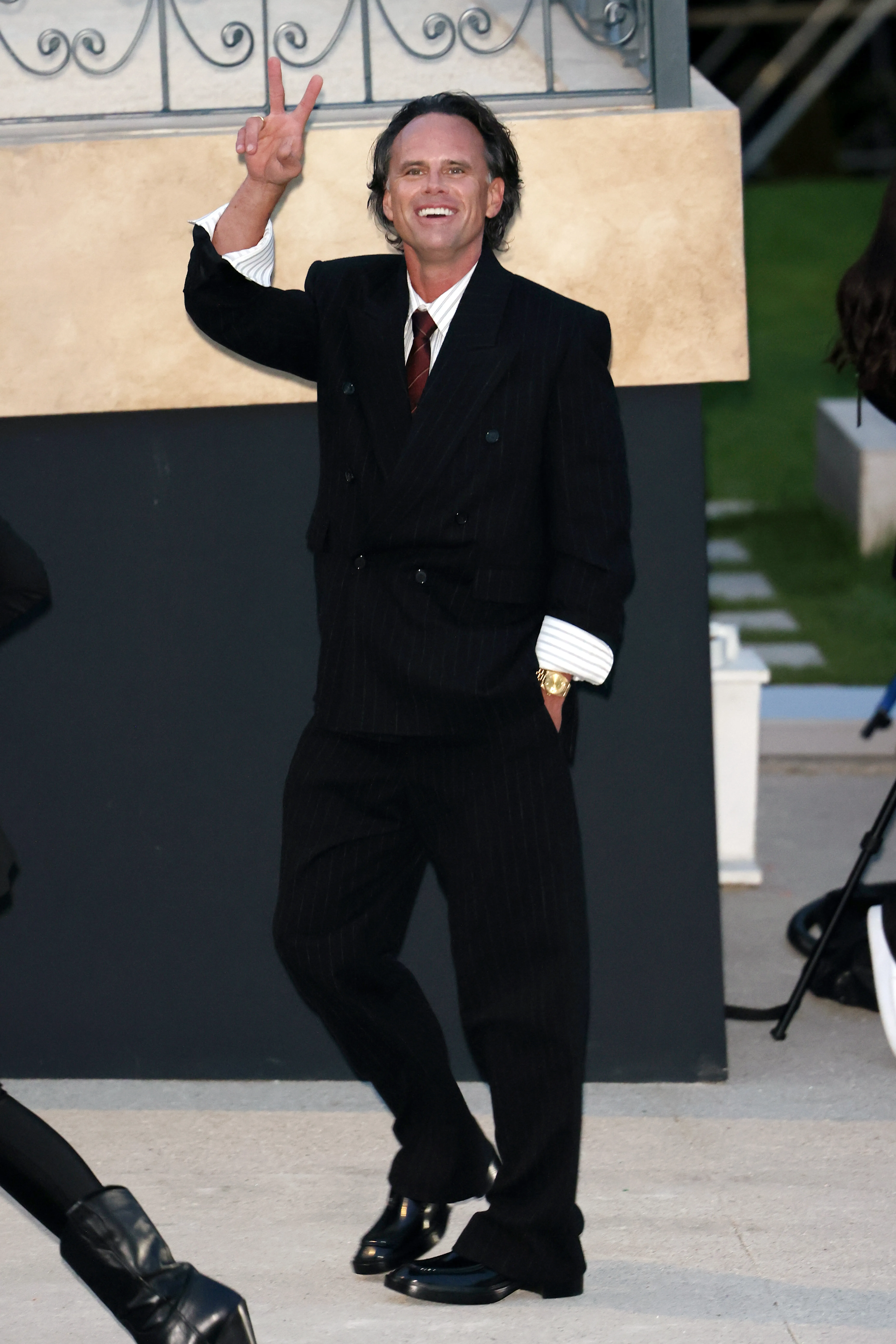 Walton Goggins in a pinstripe suit, giving a peace sign at Paris Fashion Week.