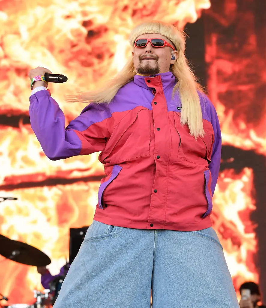 Oliver Tree performing in a red and purple jacket, baggy jeans, red sunglasses, and a blonde wig, holding a microphone.