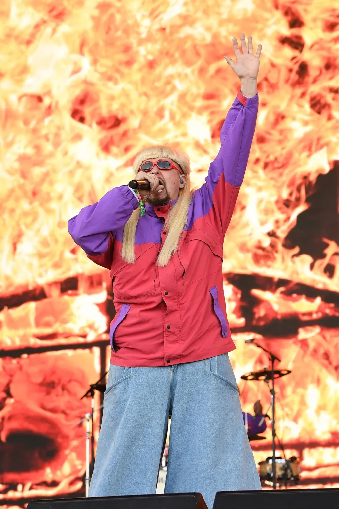 Oliver Tree performing on stage, wearing a blonde wig, red sunglasses, a red and purple jacket, and wide-legged denim jeans, with a fiery backdrop.