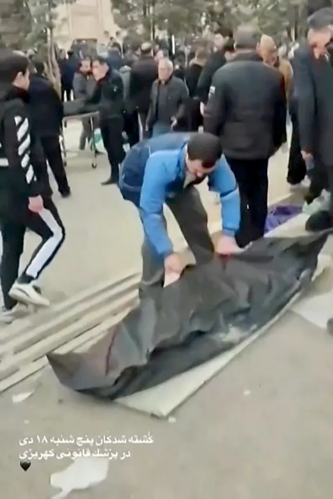 A man in a blue jacket bends over a body bag on the ground, surrounded by other body bags and people in what appears to be an outdoor medical facility.