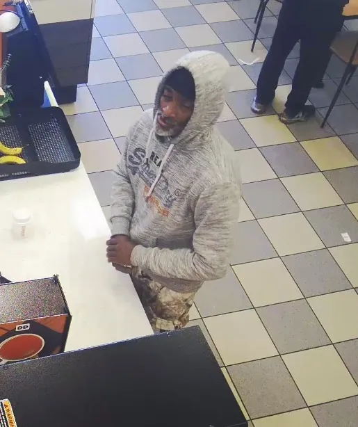 A man in a grey hoodie, dark beanie, and camouflage pants stands at a counter.