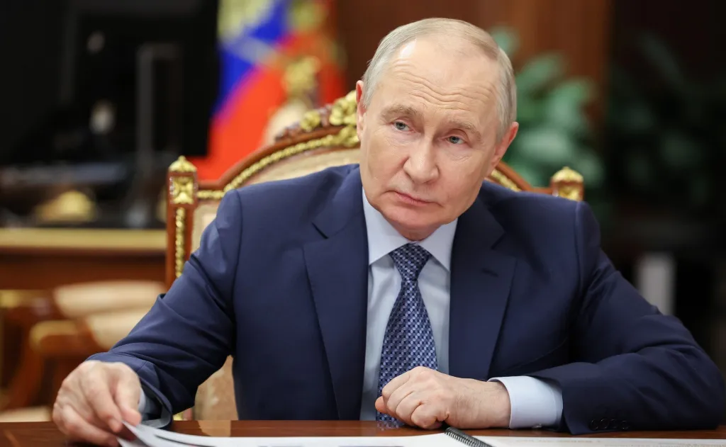 Russian President Vladimir Putin in a dark suit and patterned tie, seated at a wooden table.