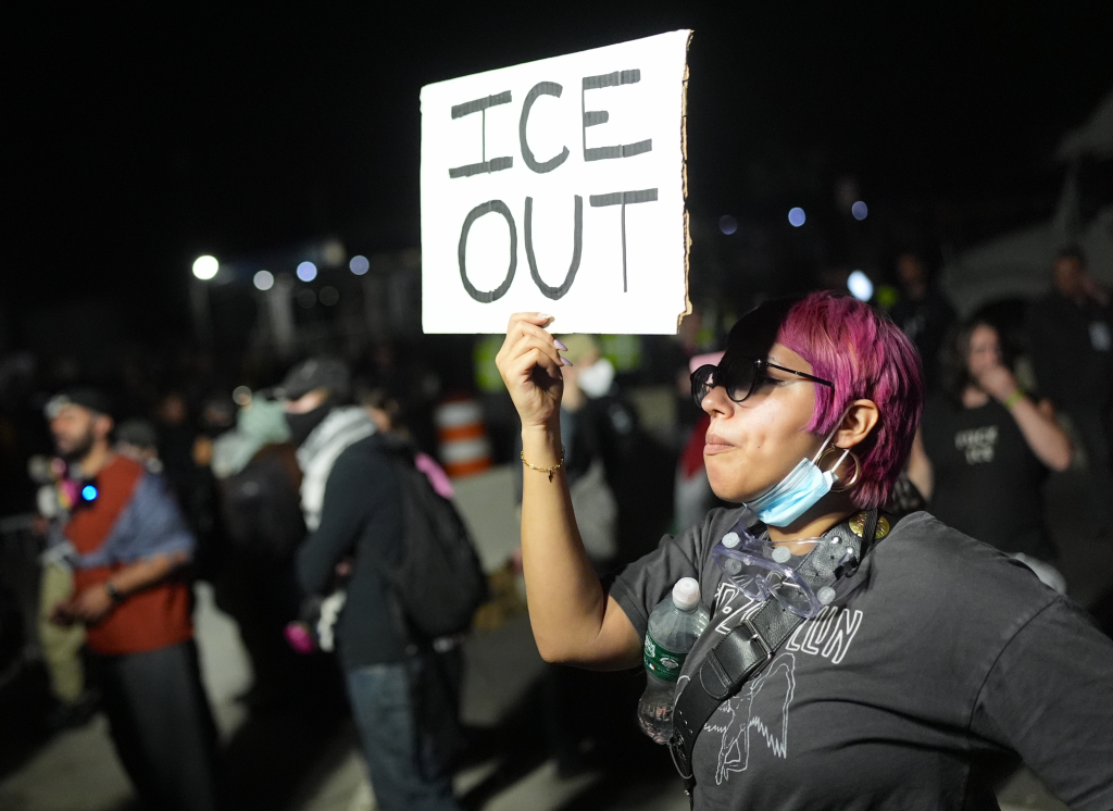 A protestor with pink hair holds a sign that reads "ICE OUT" at night.