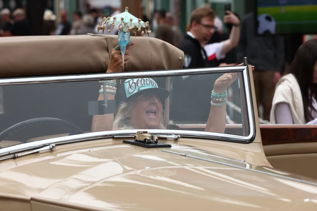 A woman in a "Beachy" cap smiles from inside a vintage car, holding a parasol decorated with seashells and a blue bird.