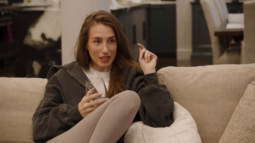 A young woman in a dark gray hoodie and light leggings sits on a light beige couch, holding a glass and gesturing with her right hand.