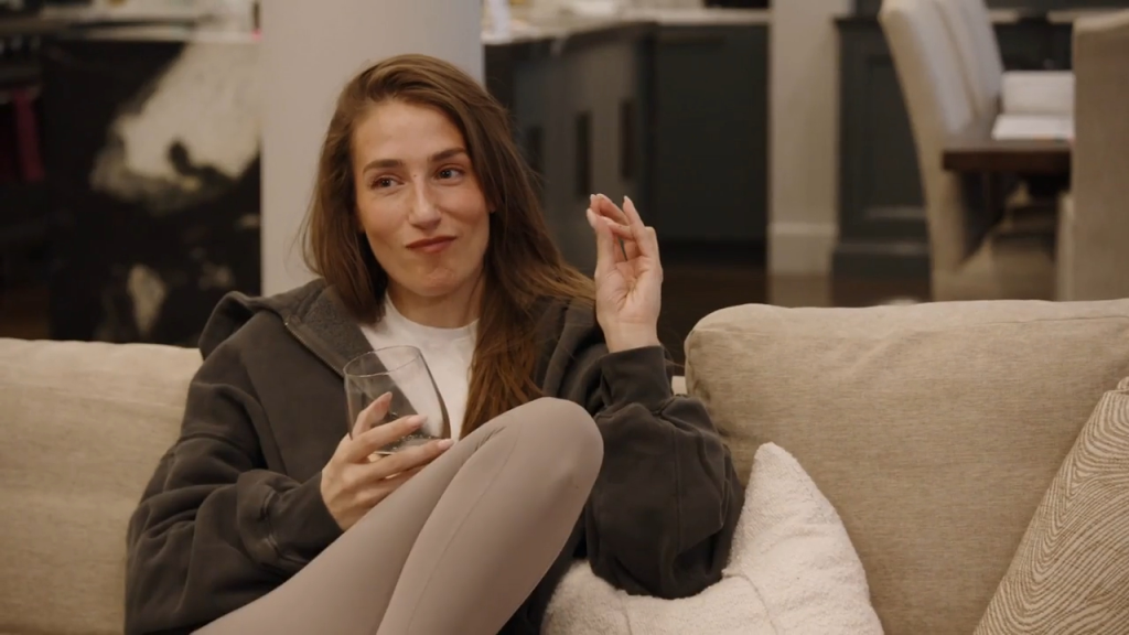A woman sitting on a couch with a glass in her hand, making a gesture with her other hand.