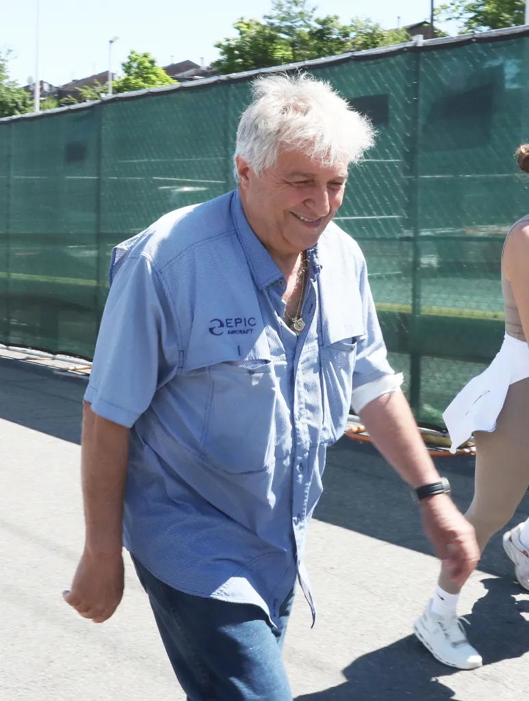 Giuseppe Oppedisano walking while smiling, wearing a light blue shirt with "EPIC AIRCRAFT" logo, dark blue jeans, and a pendant.